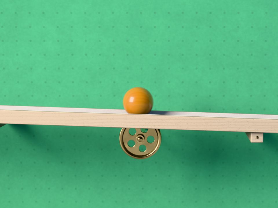 La bola rueda por un listón de haya inclinado, con una polea de latón debajo, delante de una pared verde salvia.